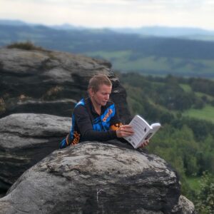Autorka Wiolka Czuryńska siedzi na skałach i czyta książkę w wersji papierowej „Zrób tylko jeden krok. Tom 1” na tle rozległego, górskiego krajobrazu.