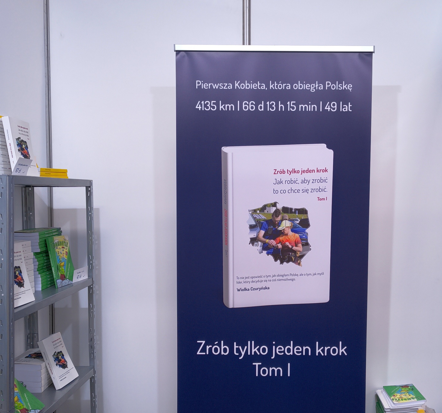 Duży granatowy baner promujący książkę Wiolki Czuryńskiej „Zrób tylko jeden krok. Tom 1” postawiony obok regału z książkami.