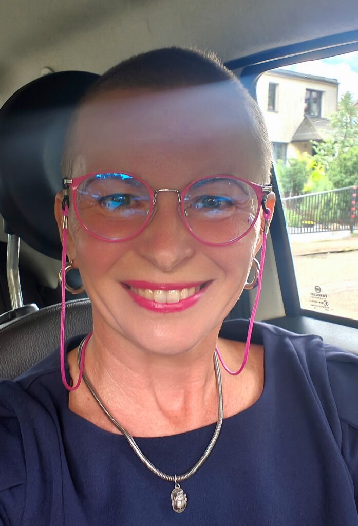 Wiolka Czuryńska, ultramaratonka, w okularach o różowych oprawkach ma bardzo krótkie włosy, granatową elegancką sukienkę, ale widać tylko popiersie. Wiolka siedzi w samochodzie na miejscu kierowcy, ma piękny szeroki uśmiech, usta mocno różowe idealnie pasujące do okularów. Na szyi gruby łańcuszek z wisiorkiem przedstawiającym skarabeusza, a na uszach grube koła o średnicy około 1,5 cm.
